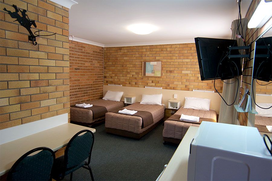 A room with three single beds with brown sheets, a TV set up, two black chairs